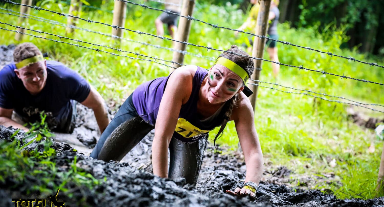 A woman in athletic gear and face paint crawls through mud under barbed wire during an outdoor obstacle race, followed by other participants. Bespoke glass solutions and the Total Warrior logo are visible in the corner.