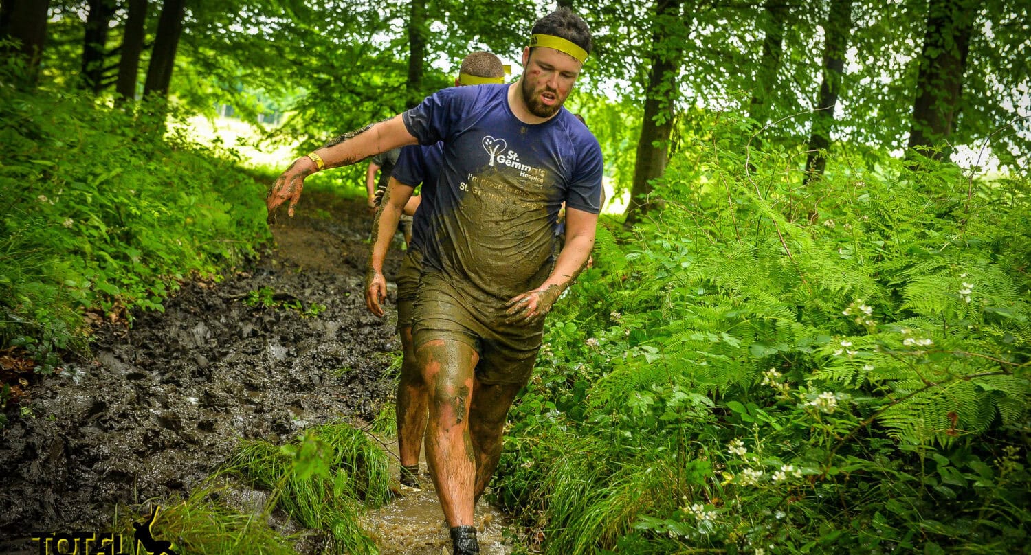 A man in a headband and muddy sportswear runs through a forest trail, splashing through thick mud like hes racing toward adventure-ready front doors. Another participant follows as lush green trees and ferns surround the path. Total Warrior logo is visible in the corner.