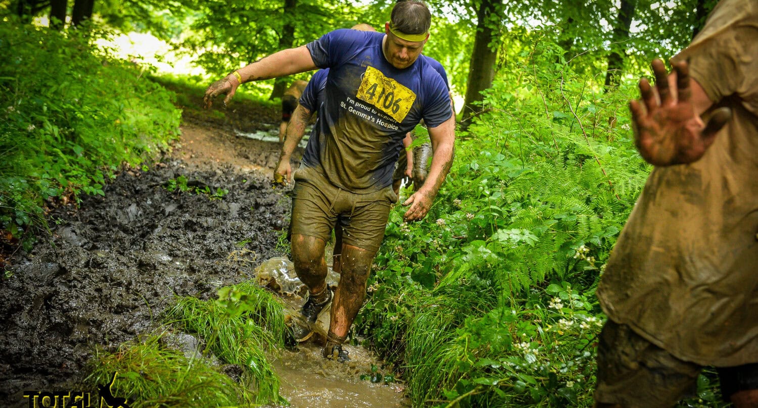 A man wearing a race bib and headband runs through a muddy forest trail during a Total Warrior obstacle event, splashing water and mud—much like sliding doors opening to new challenges. Another muddy participant is partially visible on the right.