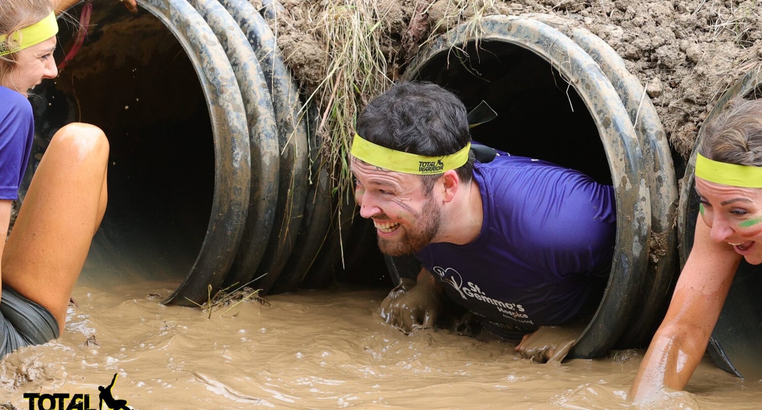 Three people wearing yellow headbands crawl through muddy pipes during an obstacle course, smiling and covered in mud. Glass roofing shelters part of the run, while the “Total Warrior” logo is visible in the bottom left corner.