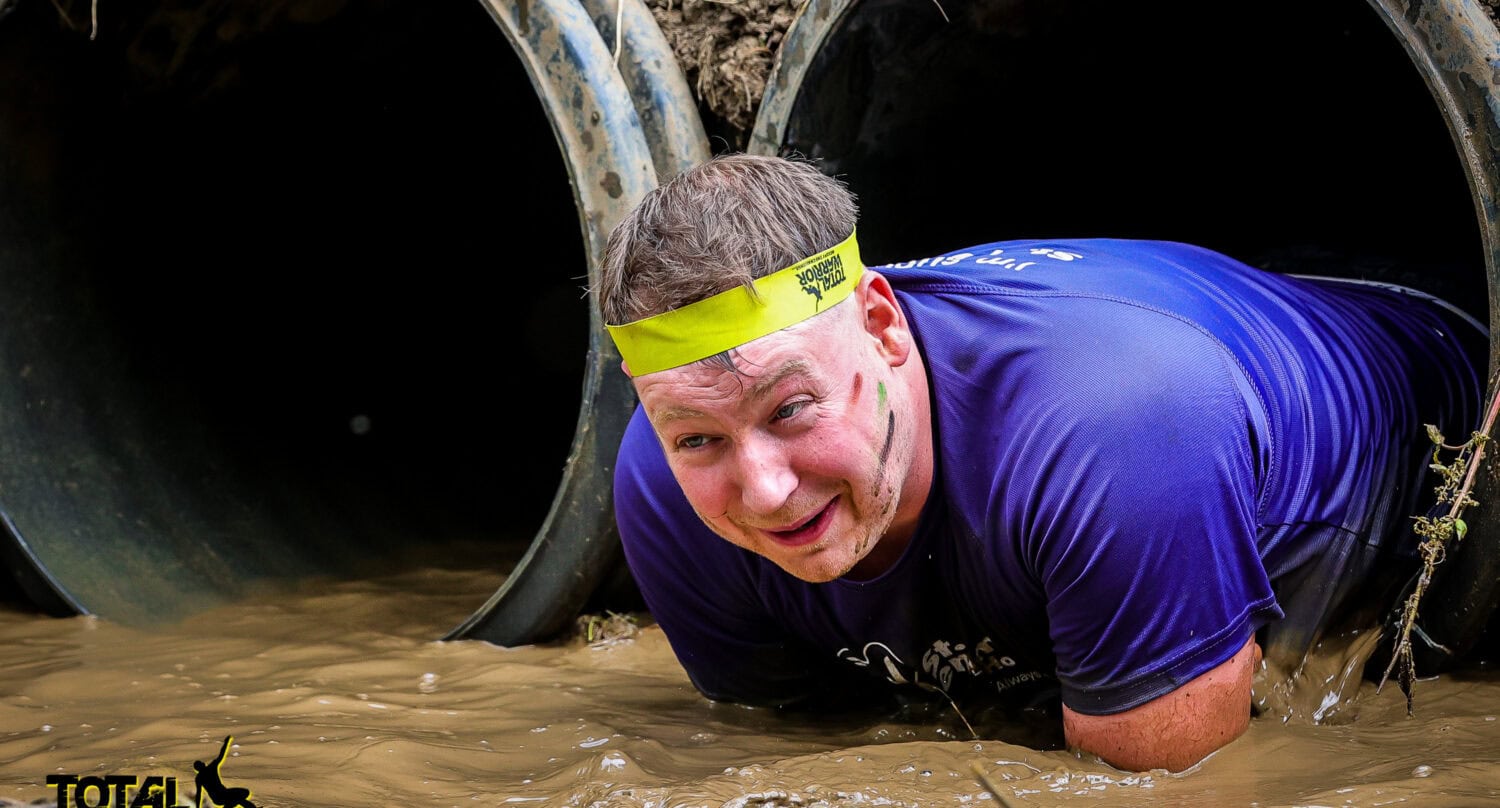 A man wearing a yellow headband and blue shirt crawls through a muddy tunnel during an obstacle race, with muddy water on the ground. The Total Warrior logo appears in the corner, reflecting his drive for bespoke glass solutions in every challenge.