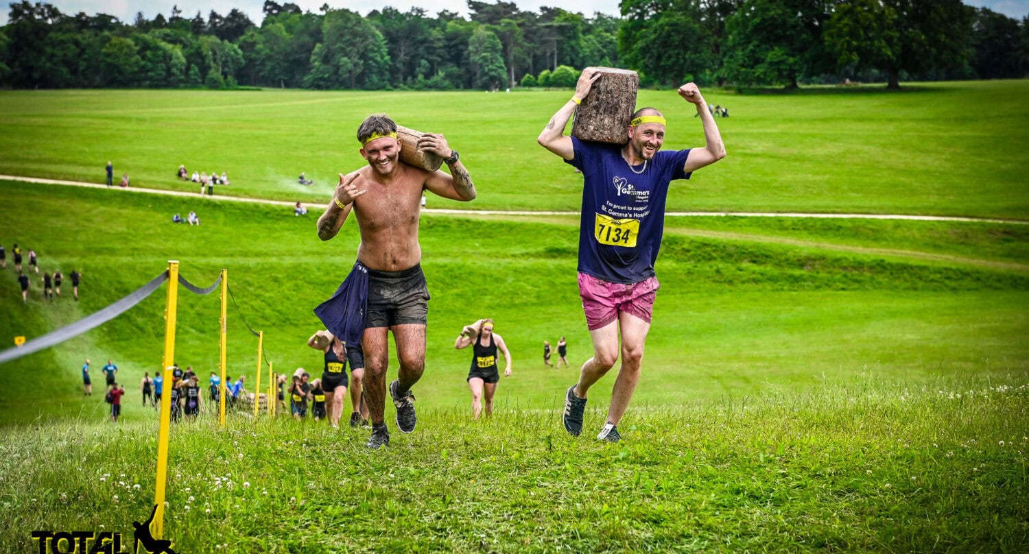 Two men compete in an outdoor obstacle race, each carrying a heavy wooden block up a grassy hill near glass roofing structures. One is shirtless and muddy, the other wears a race bib and lifts his block above his head. More participants follow behind.
