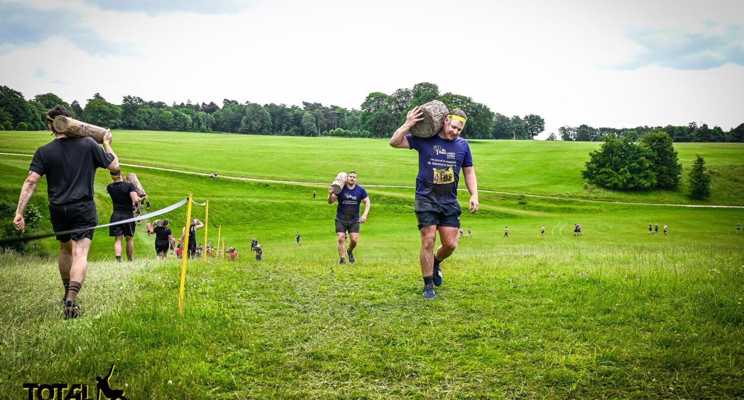Participants in athletic clothing carry large logs on their shoulders while running up a grassy hill, passing stacks of front doors as part of the outdoor Total Warrior obstacle course. Trees and a cloudy sky provide a dramatic backdrop.