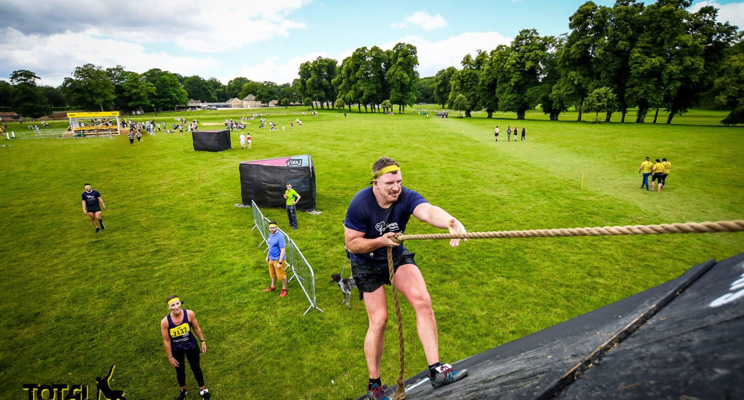 A man in athletic gear climbs a steep obstacle using a rope during an outdoor Total Warrior event, with other participants and green fields visible in the background, reminiscent of the teamwork needed to move heavy sliding doors into place.