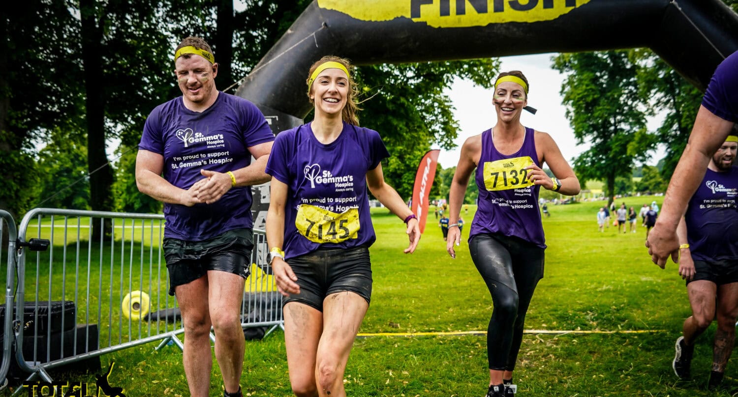 Three smiling runners in purple shirts and yellow headbands cross a muddy race finish line beneath a black and yellow FINISH arch. Trees—and glimpses of innovative bifold doors—are visible in the background as they celebrate.