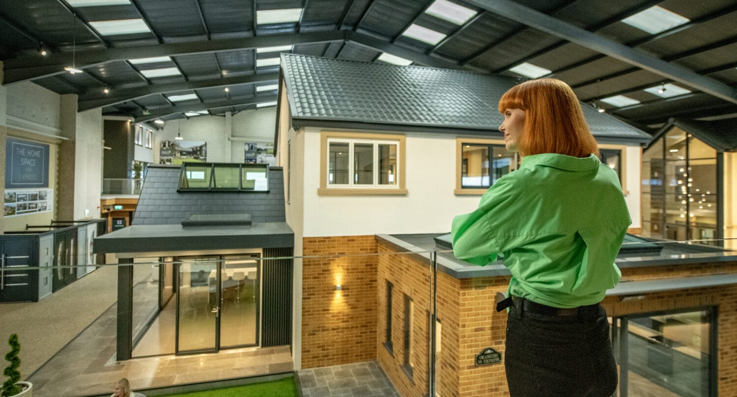A woman with red hair wearing a bright green shirt stands on a balcony overlooking a modern indoor exhibition space featuring model homes with glass roofing and people seated at a table below.