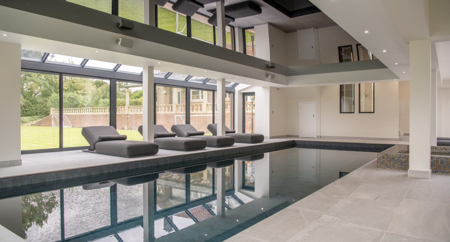 Modern indoor swimming pool room with large windows, poolside loungers, light-colored tile flooring, and a reflective ceiling. Green lawn and trees are visible outside through bespoke glass sliding doors.