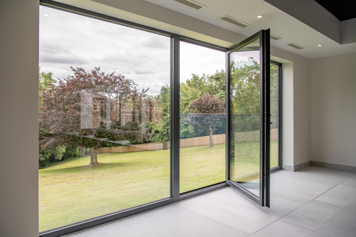A modern room with large floor-to-ceiling sliding doors, one of which is open, overlooks a grassy lawn with several trees and a wooden fence under a cloudy sky.