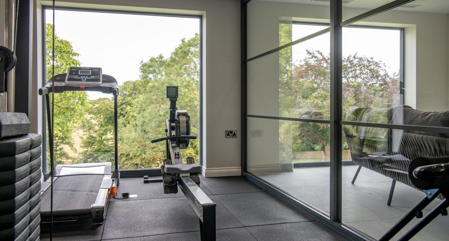 A modern home gym with a treadmill and rowing machine, featuring large windows overlooking green trees and a bespoke glass wall separating the gym from a living area with a sofa.