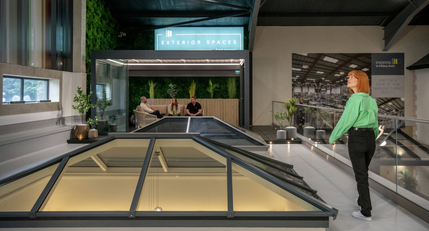 A woman in a green shirt stands indoors near large, angled glass roofing skylights. In the background, two people sit in a lounge area labeled Exterior Spaces, surrounded by greenery and modern decor.