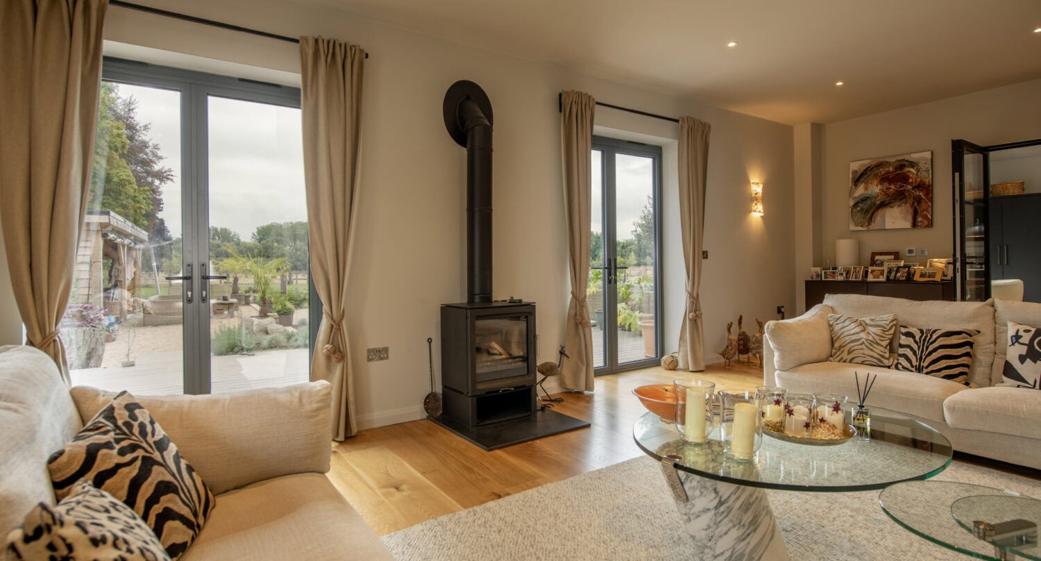 A cozy living room with light-colored sofas, patterned cushions, a wood-burning stove, glass coffee tables, and large windows with beige curtains overlooking a garden patio through elegant sliding doors.