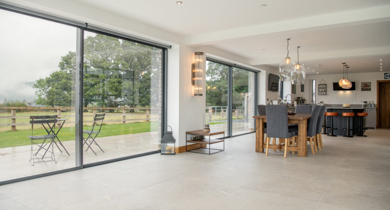 Modern open-plan dining area with large bifold doors leading to a patio, outdoor seating, and a grassy yard with a wooden fence and trees. Neutral-toned flooring and contemporary lighting are featured inside.