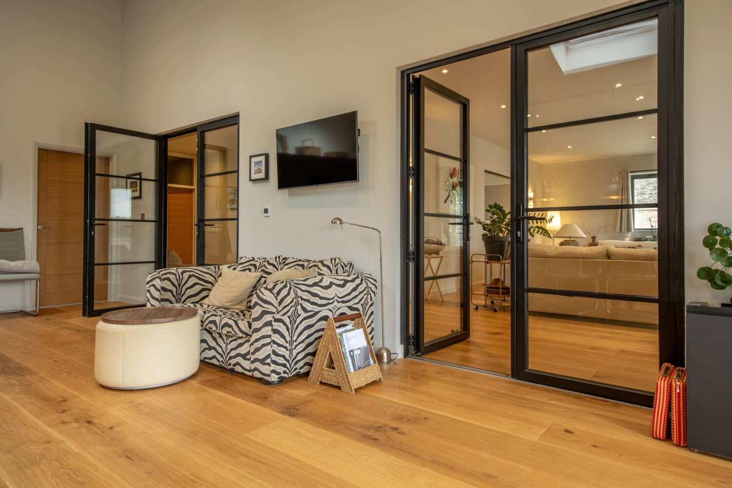 Modern living room with wood floors, a zebra-print sofa, a round ottoman, wall-mounted TV, and large bespoke glass bifold doors leading to another stylishly furnished room. A rattan basket with magazines sits by the sofa.