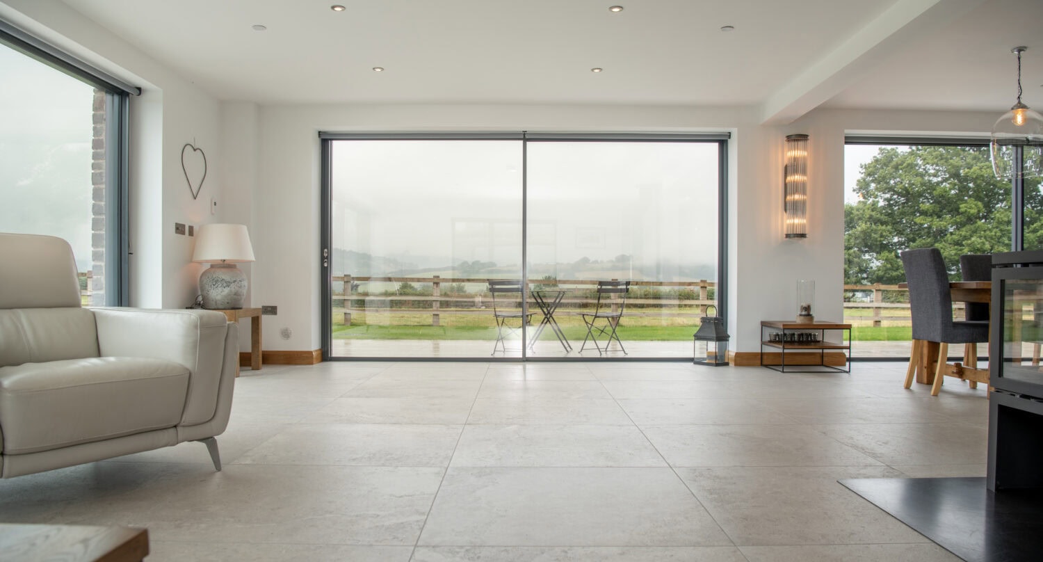 Modern living and dining area with large sliding glass doors and bifold doors overlooking a patio and green landscape; light-colored tiles, minimal furnishings, and natural light filling the space.