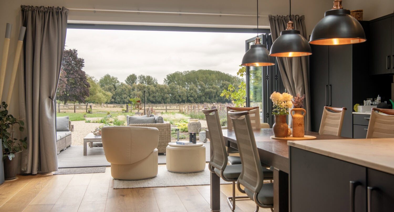 Modern open-plan kitchen and dining area with pendant lights, leading to a cozy patio with outdoor seating; large sliding doors reveal a scenic garden and trees beyond.
