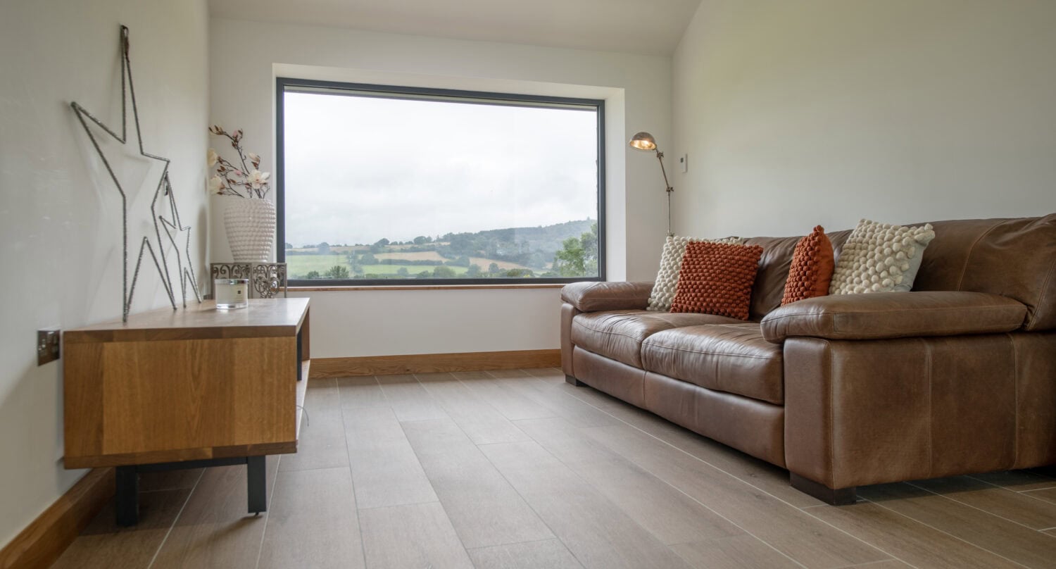 A minimalist living room with a large picture window and sliding doors framing a scenic countryside view, brown leather sofa with patterned cushions, wooden cabinet, and neutral decor with star-shaped wall art.