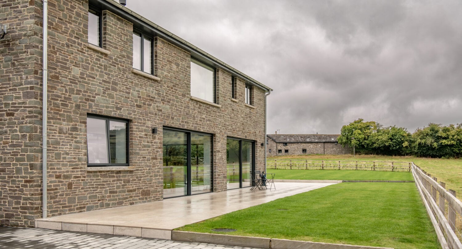 A modern stone house with large windows and sliding doors, featuring glass roofing that bathes the patio and green lawn in natural light, set under a cloudy sky with distant trees and a wooden fence.