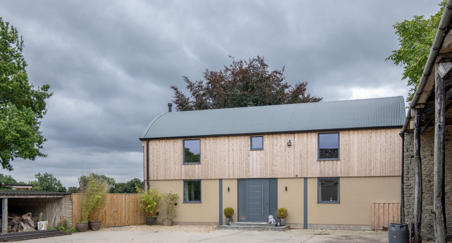 Modern two-story house with a curved metal roof, wooden and beige walls, large bifold doors, and several windows. The house is surrounded by a paved courtyard, plants, and trees under a cloudy sky.