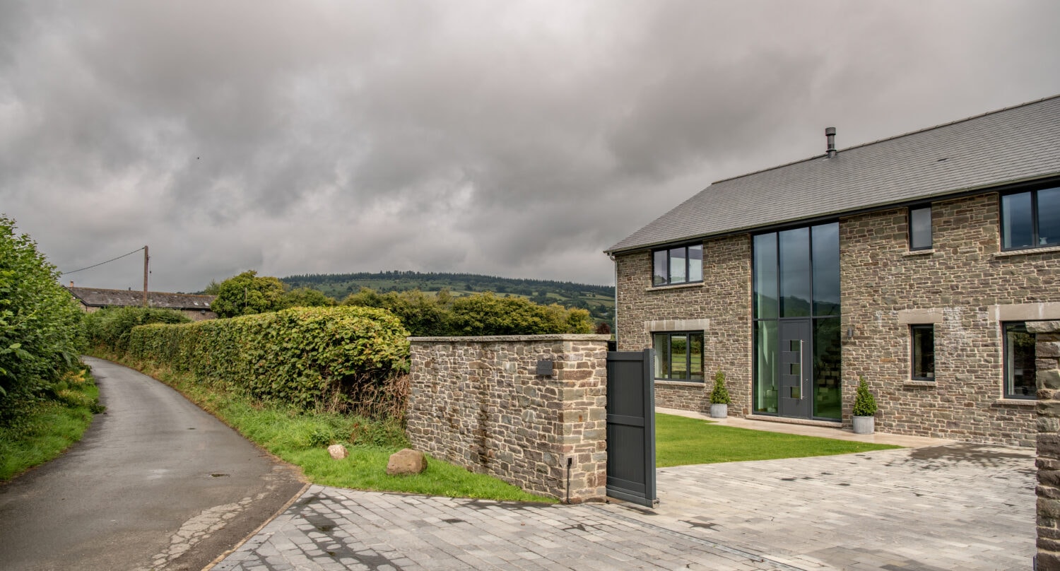 A modern stone house with large glass windows, stylish bifold doors, a paved driveway, and a stone wall, set in a rural landscape with a narrow road, green hedges, and cloudy skies.