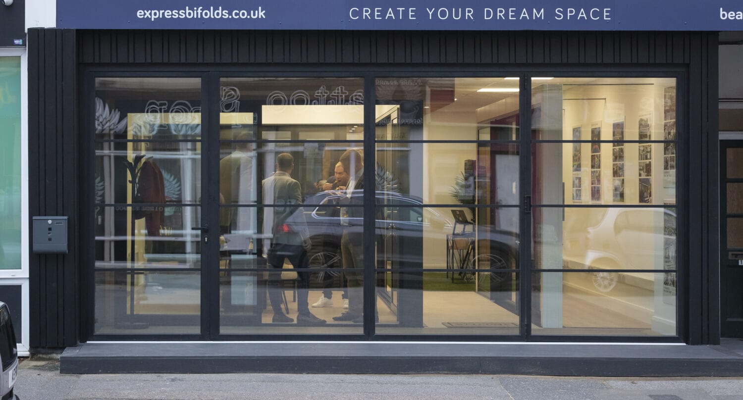 A modern storefront with large glass windows shows several people talking inside. The sign above reads “CREATE YOUR DREAM SPACE” and advertises sliding doors, bifold doors, and stylish glass roofing options.