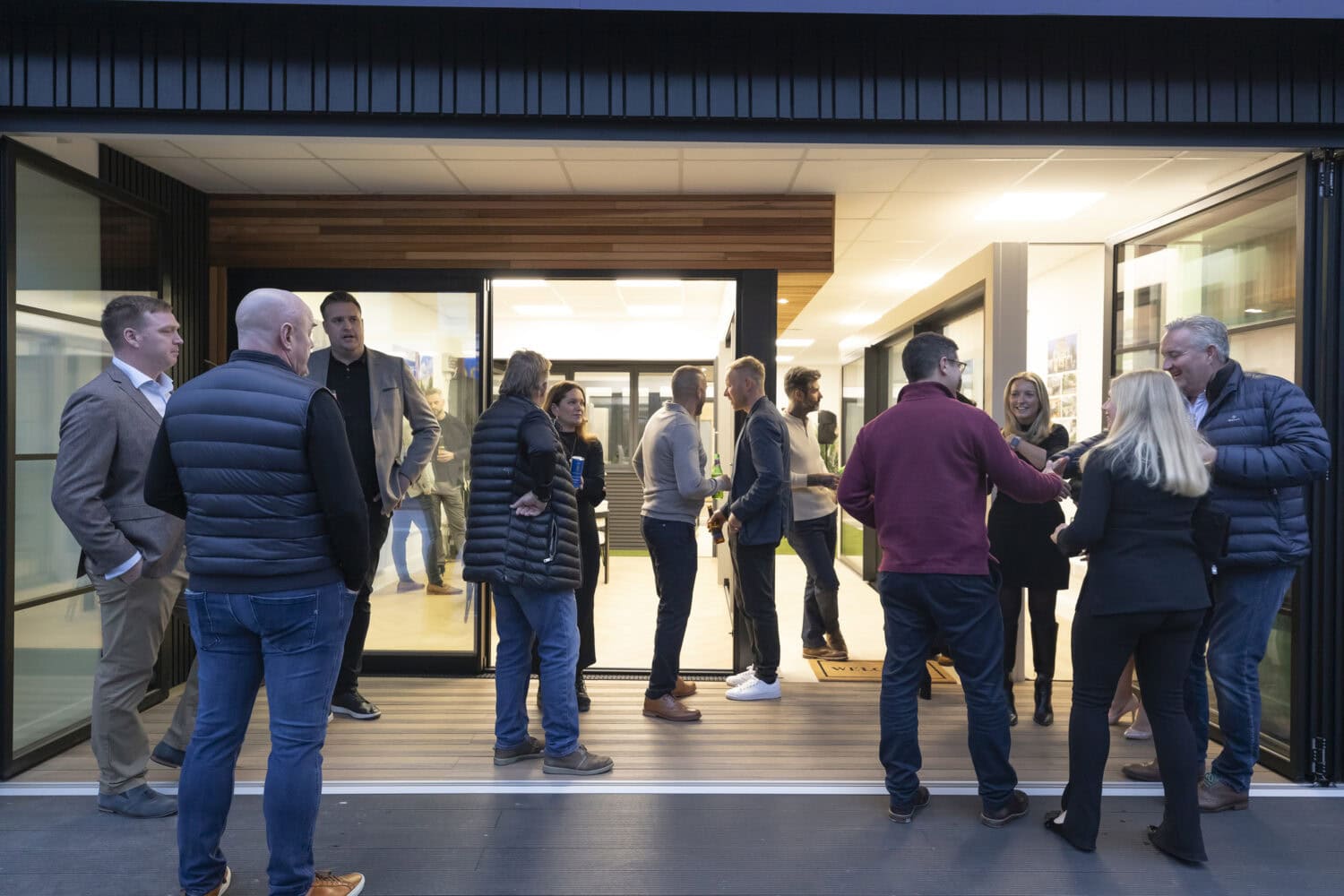 A group of people stand and chat outside and inside a modern building with bespoke glass solutions, including sleek glass doors. Some are shaking hands, while others engage in conversation in this professional or social setting.