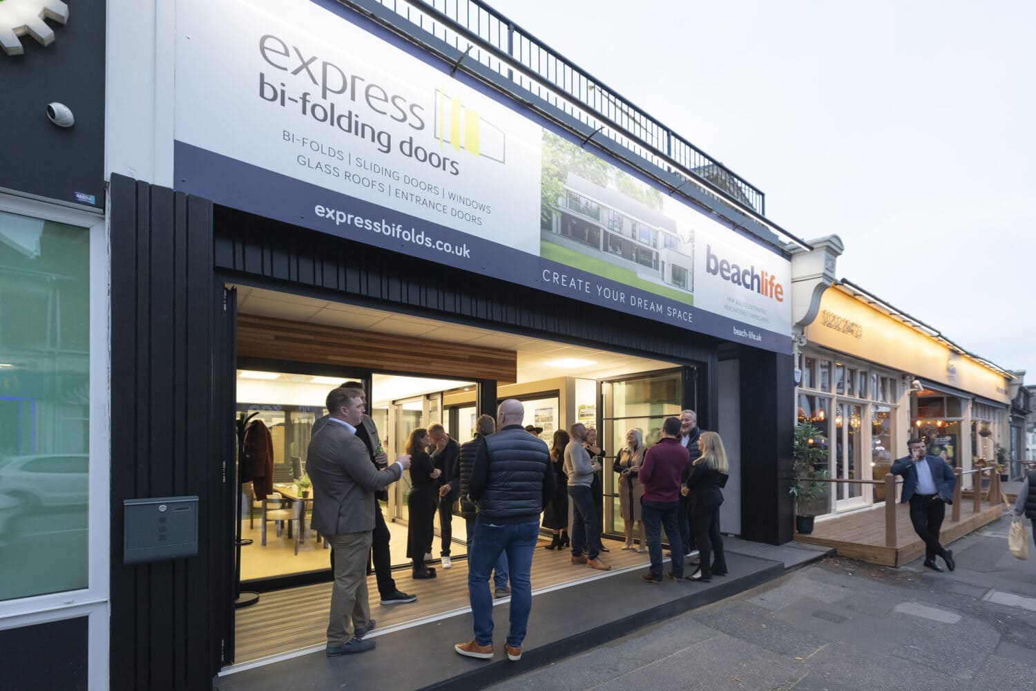 A group of people converse outside and inside a modern storefront for Express Bi-Folding Doors, where sleek bifold doors are on display. Next door, there’s a “beachlife” sign. The scene is set in the evening on a busy street.