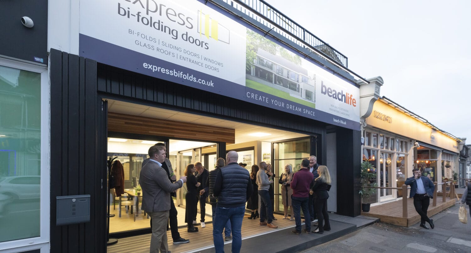 A group of people converse outside and inside a modern storefront for Express Bi-Folding Doors, where sleek bifold doors are on display. Next door, there’s a “beachlife” sign. The scene is set in the evening on a busy street.