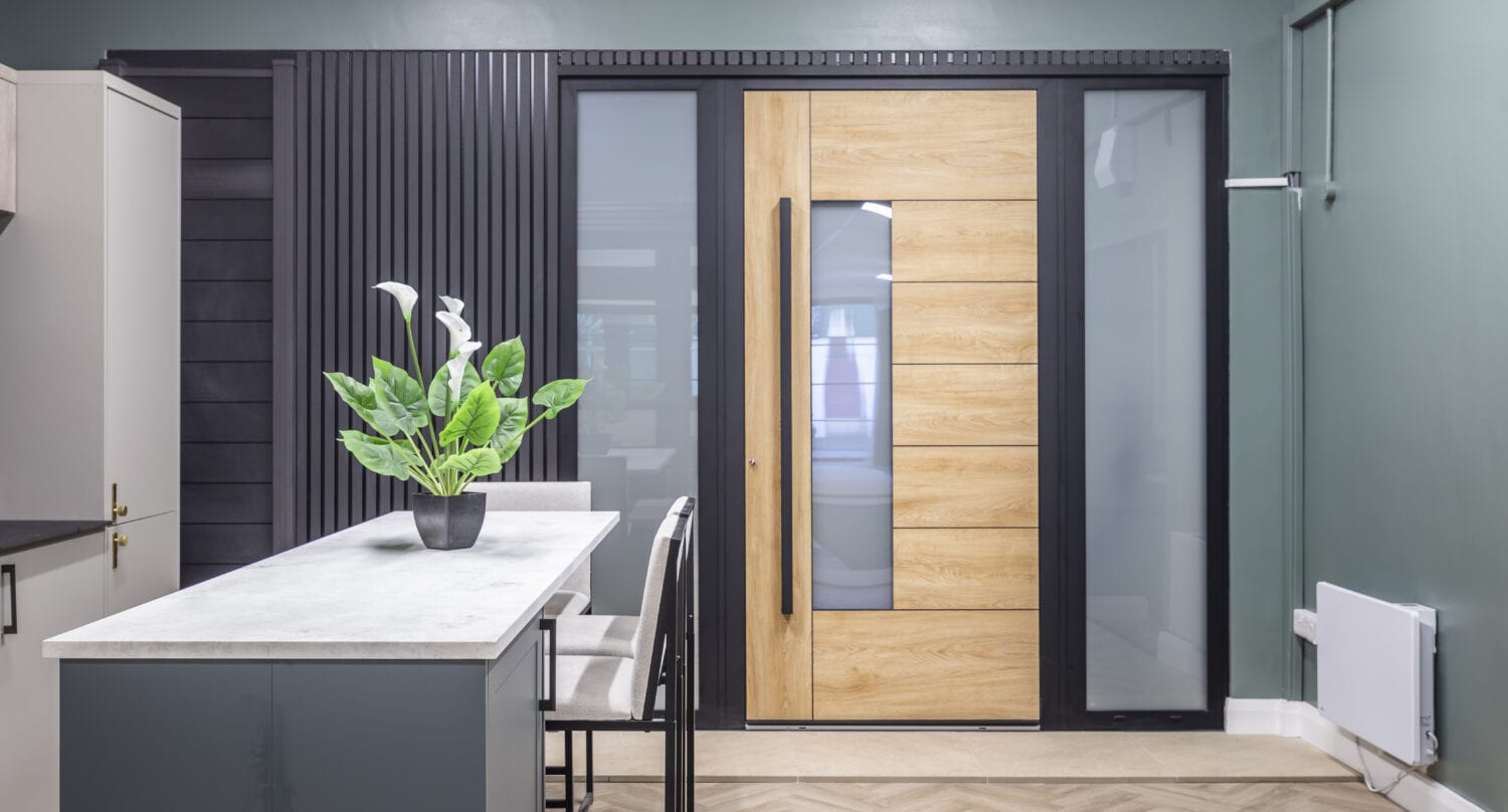 Modern kitchen with a green accent wall, a light wood door with frosted glass panels, bespoke glass solutions on the marble-topped island, two bar stools, and a potted plant on the countertop.