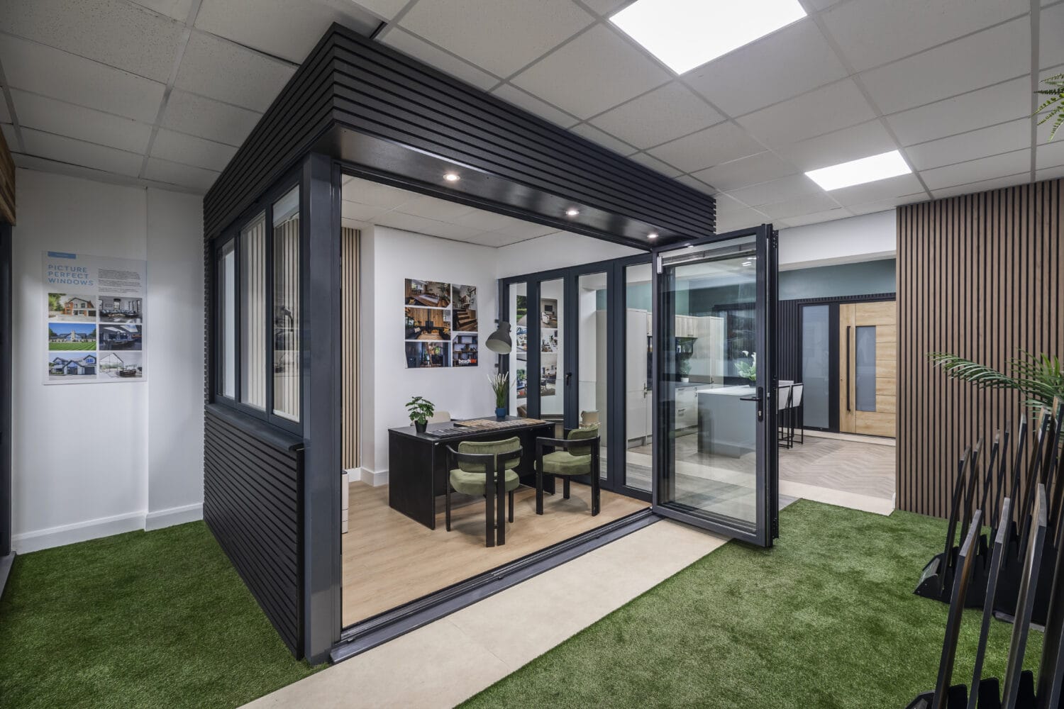 A modern office space with glass walls, black frame, and an open door. Inside are a desk, chairs, and wall art. The floor combines artificial grass and wood, while sliding doors and ceiling lights create a bright, airy atmosphere.