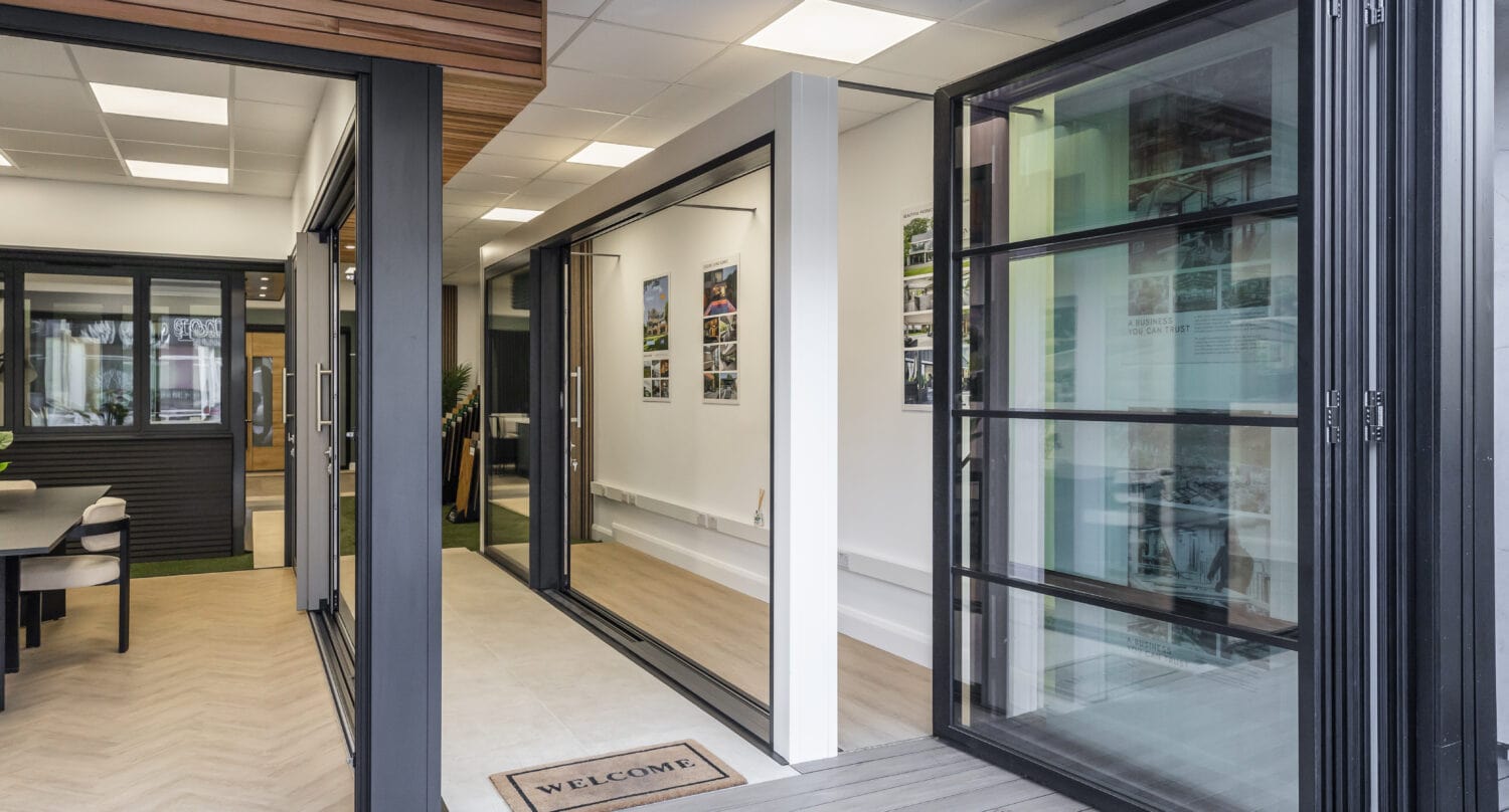 A modern interior showroom with large bifold doors, light wood flooring, white walls, and a “Welcome” doormat at the entrance. Framed pictures hang on the wall inside.
