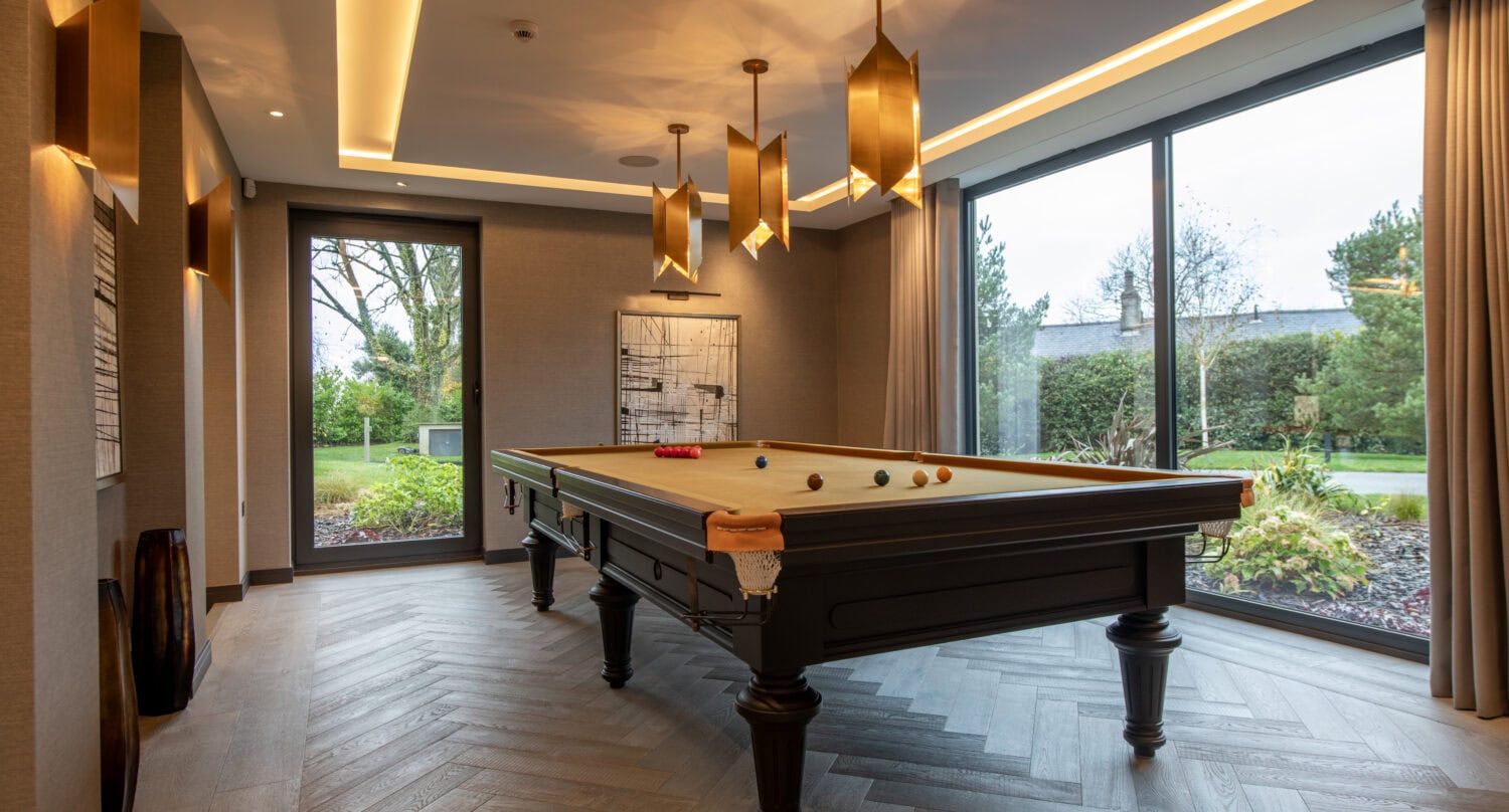 A modern room with large windows and bifold doors features a billiards table set for a game, pendant lights hanging above, wood flooring, and views of a garden outside.