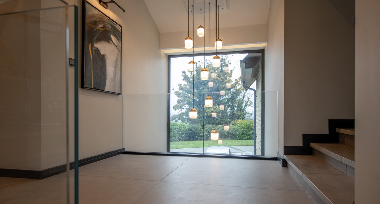 A modern interior space with hanging pendant lights, a large window overlooking greenery, sliding doors near the staircase with dark steps, and contemporary wall art on the left.