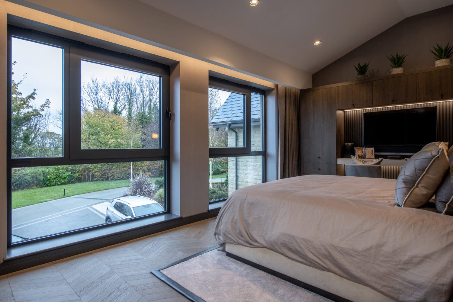Modern bedroom with large windows and sliding doors overlooking a driveway and trees, featuring a neatly made bed, wood flooring, a wall-mounted TV, and potted plants above built-in shelves. Natural light fills the room.