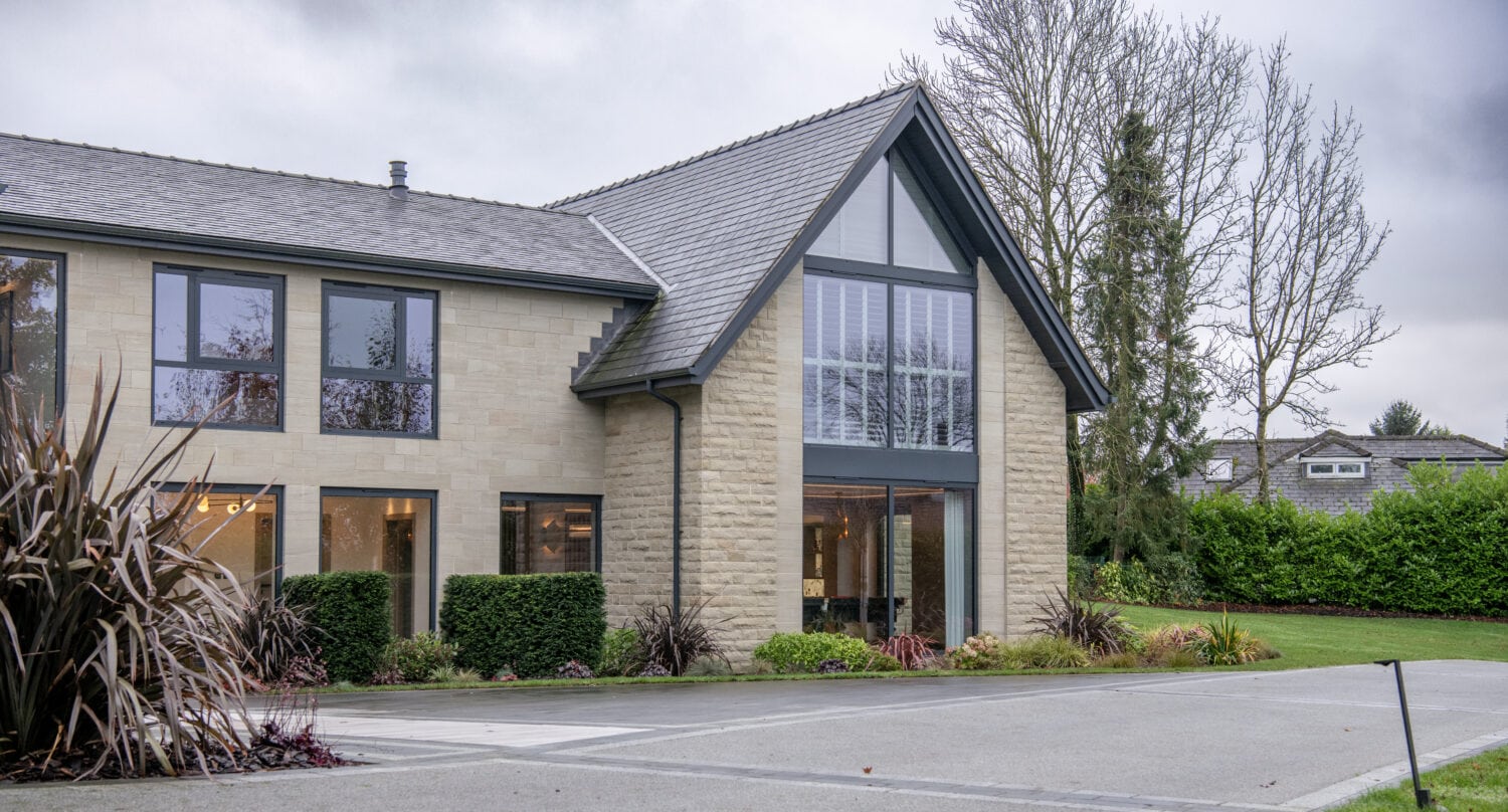 Modern two-story house with large windows, stone exterior, and triangular roof, featuring bespoke glass solutions. Surrounded by neatly trimmed bushes and trees on a cloudy day, a driveway leads up to the entrance.