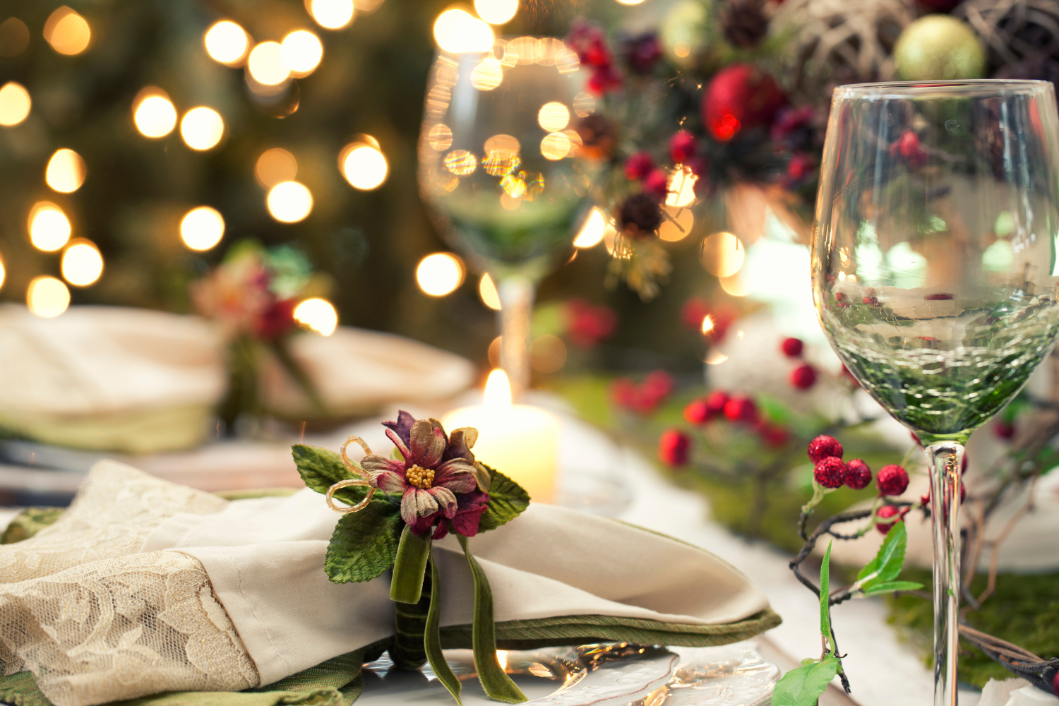 A festive holiday table setting with elegant napkins, floral napkin rings, wine glasses, and decorative berries shines softly by candlelight, with blurred golden lights framed by stunning bifold doors in the background.