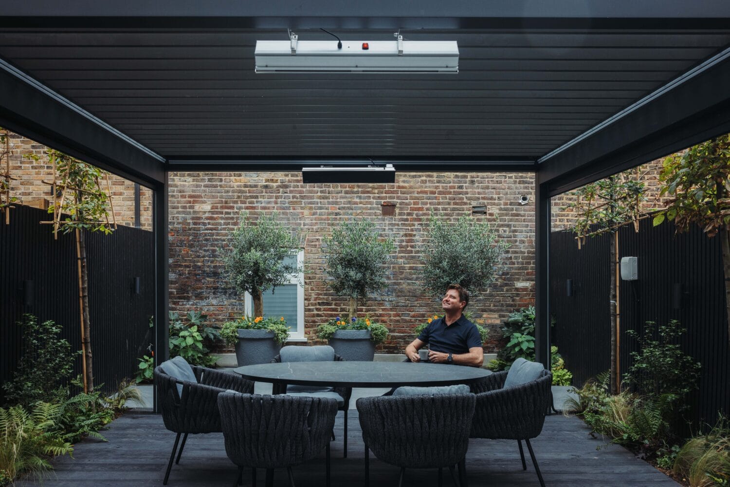 A man sits alone at a round table with six chairs in a covered outdoor patio, surrounded by potted plants and brick walls. He smiles and looks up beneath the glass roofing, enjoying the warmth from a ceiling-mounted heater.