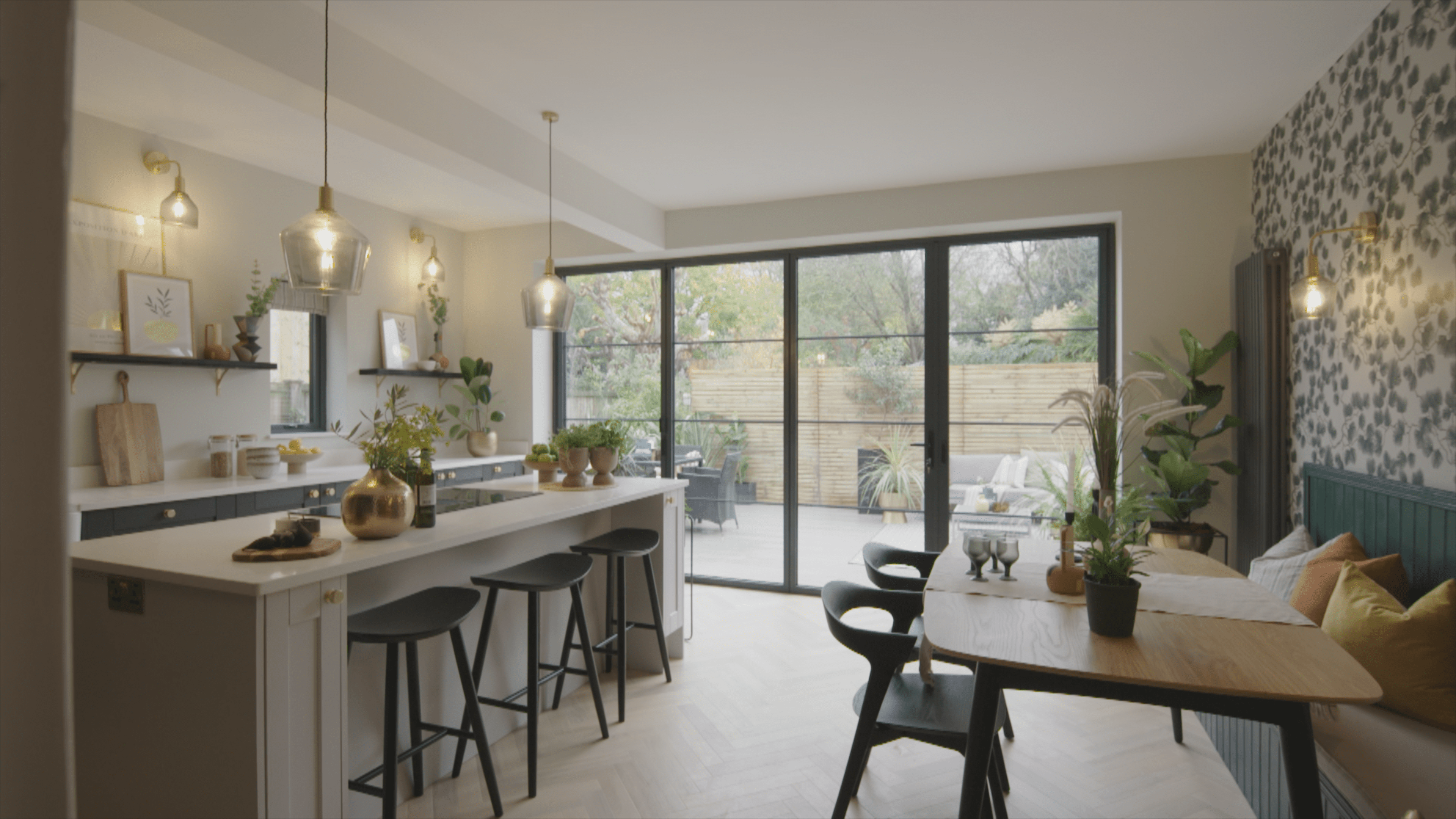 Modern kitchen and dining area with a large island, stools, pendant lights, and a wooden table. Bespoke glass solutions like sliding doors open to a patio with plants, letting in natural light. The space features greenery and contemporary decor.