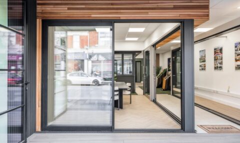 Modern office with large bifold doors, wood and metal accents, visible meeting table and chairs, wall displays, and a “Welcome” doormat. A street with parked cars is seen through the window.