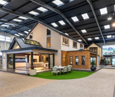 A modern, full-size house display inside a large showroom with high ceilings, featuring an open kitchen, dining area, contemporary design elements, and stunning bifold doors that open to artificial grass outside the house.
