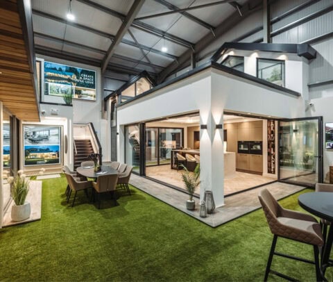 Modern showroom with artificial grass flooring, showcasing a stylish home interior with large glass walls, bifold doors, open dining and kitchen areas, potted plants, and contemporary furniture under high industrial ceilings.