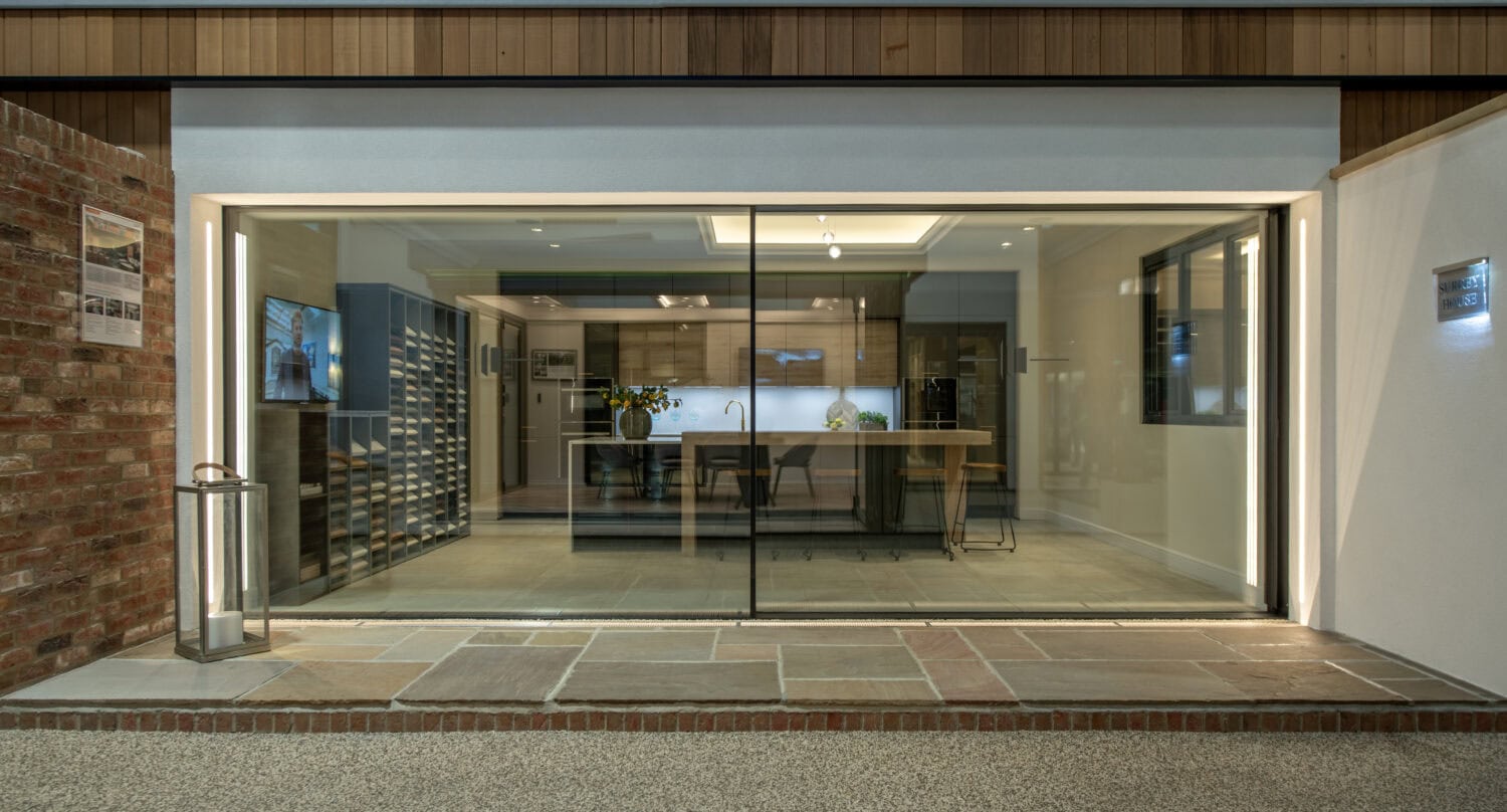 A modern kitchen showroom with large glass windows, bespoke glass solutions, sleek cabinetry, a central island, minimalist decor, and bright interior lighting, viewed from outside on a paved pathway.
