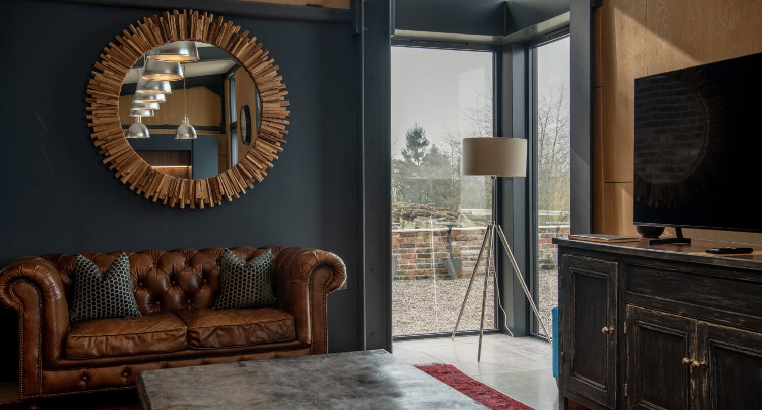 A cozy living room with a brown leather sofa, a round sunburst mirror on a dark wall, bifold doors by large corner windows, a black cabinet with a TV, and a red rug on the floor.