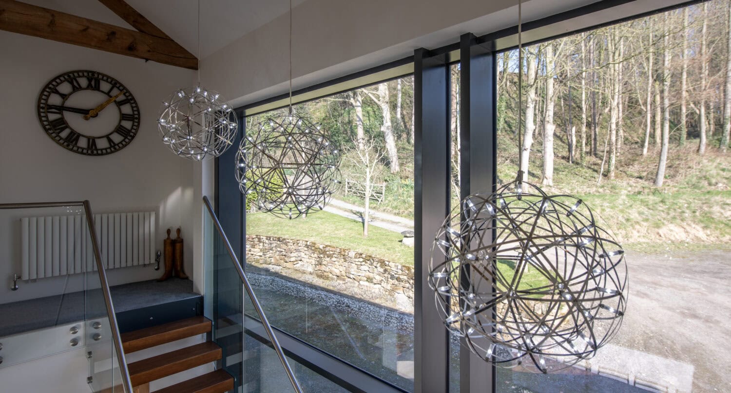 Modern stairwell with large windows showcasing a wooded area outside, three metallic pendant lights, a wooden clock on the wall, and wooden stairs with bespoke glass solutions in the railing for a stylish, open feel.