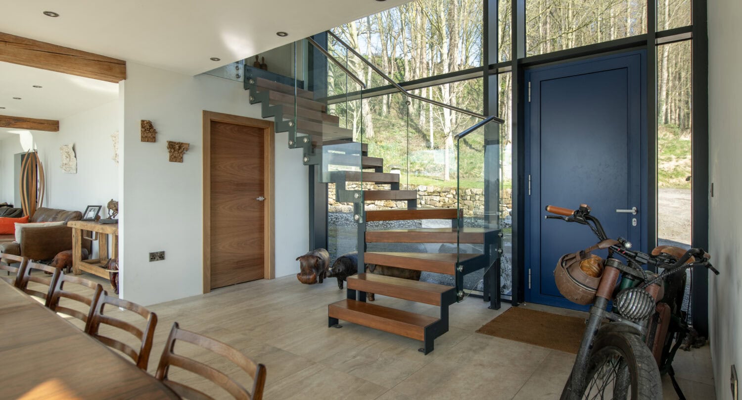 Modern entryway with blue front doors, wooden steps with glass railing leading upstairs, wooden dining table and chairs, and a bicycle with a helmet leaning against the wall. Large windows and glass roofing showcase trees outside.