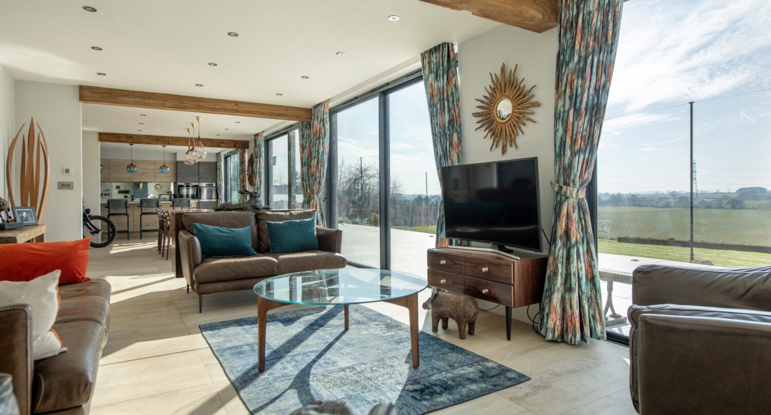 Modern living room with large windows, brown leather sofas, blue accents, sliding doors, a sunburst mirror, TV on a wooden stand, glass coffee table, and scenic countryside views. Bright, airy, and stylishly decorated.