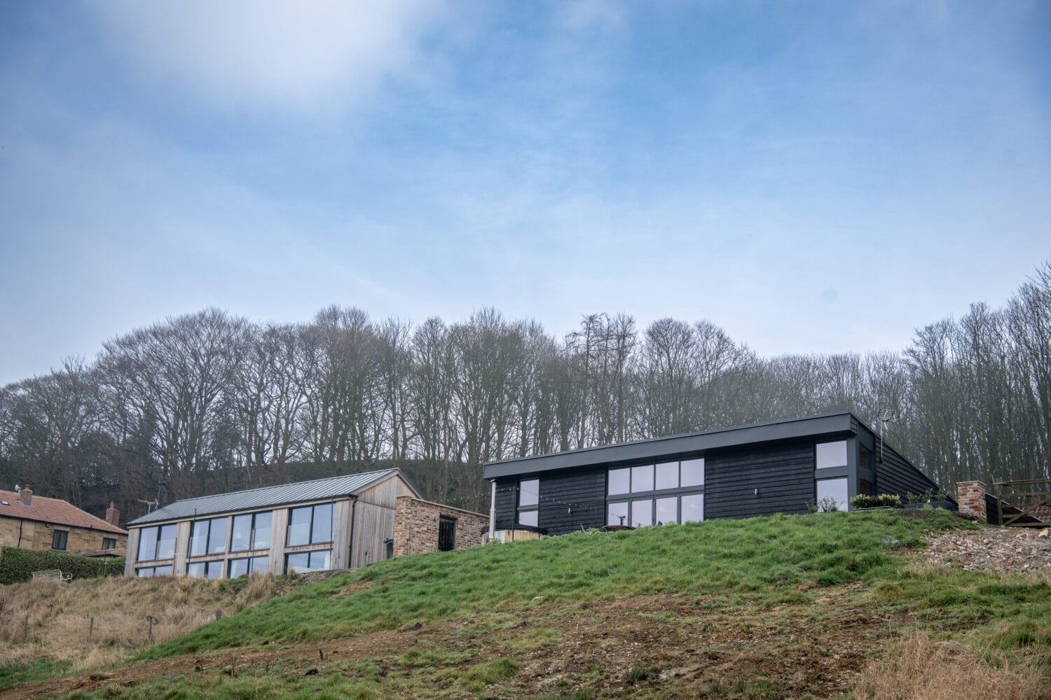 Two modern houses with large windows and sleek bifold doors sit on a grassy hillside, framed by leafless trees under a blue sky.