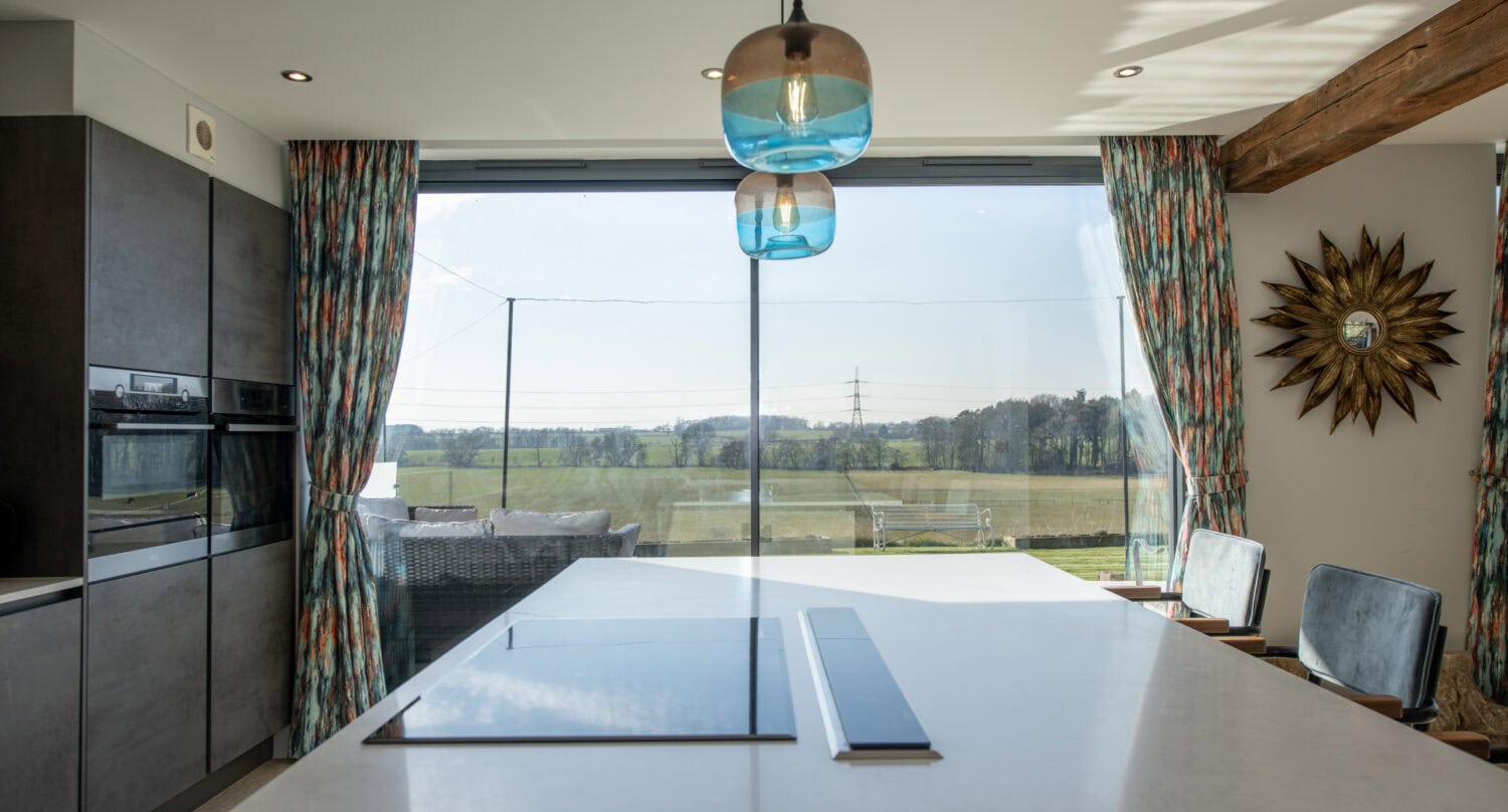 Modern kitchen with a white island countertop, built-in ovens, colorful patterned curtains, and large bifold doors overlooking a scenic outdoor landscape. Blue hanging lights and a sunburst wall decor brighten the space.