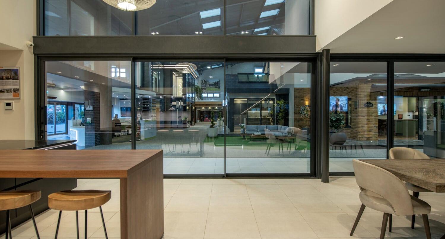 Modern interior with wooden bar stools, a table, and chairs in the foreground. Large bespoke glass solutions for doors reveal a spacious, bright courtyard with seating and greenery outside. The area feels open, stylish, and inviting.