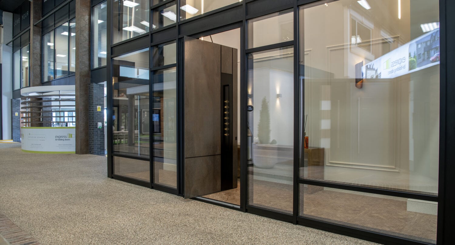 Modern glass office entrance with a brown front door, transparent walls, and carpeted floor in a building lobby. Reflections of ceiling lights are visible on the glass. A reception desk is seen in the background.