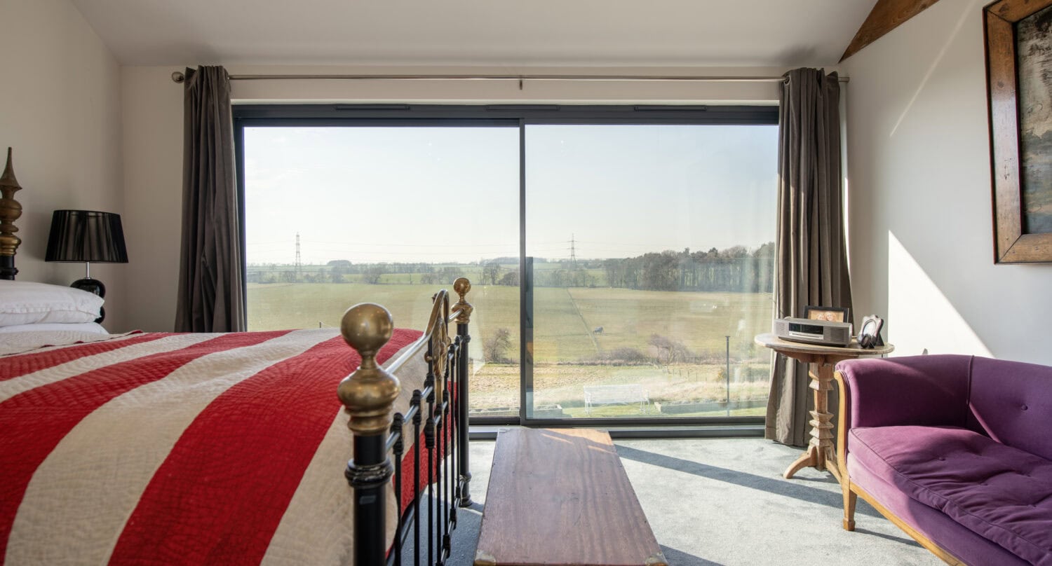A bright bedroom with a brass bed covered by a red and white striped quilt, a wooden chest at the foot, a purple sofa, and large bespoke glass sliding doors offering views of open fields and distant trees.